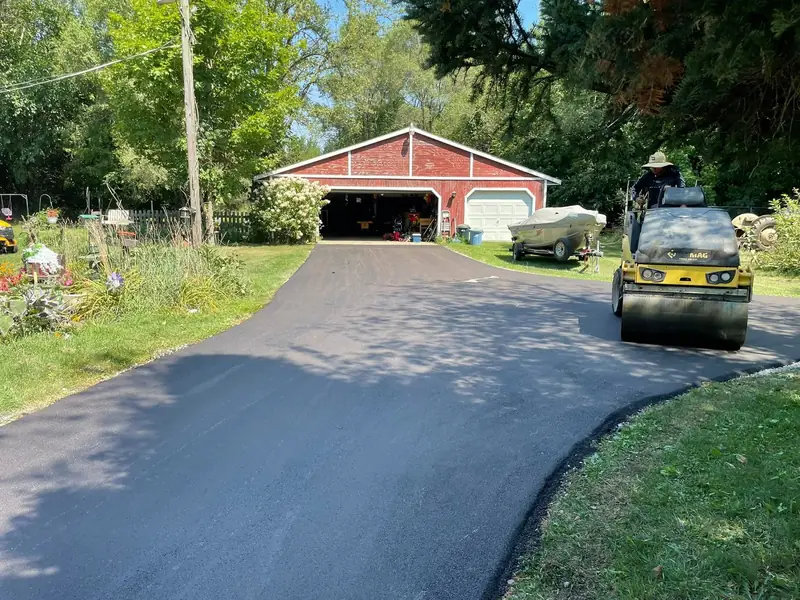 Roller finishing a new asphalt driveway that leads to a red barn garage