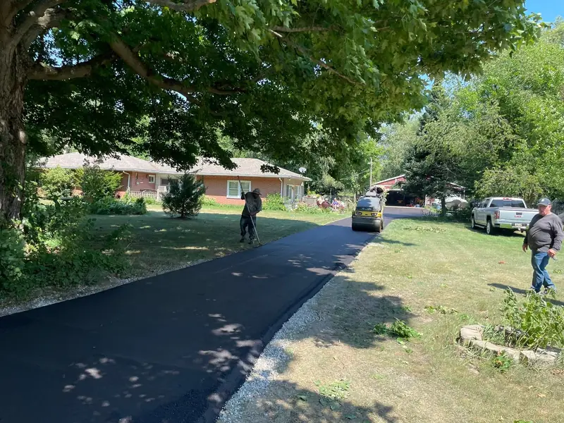 Crew finishing a new driveway under a big shade tree while the roller works toward the street