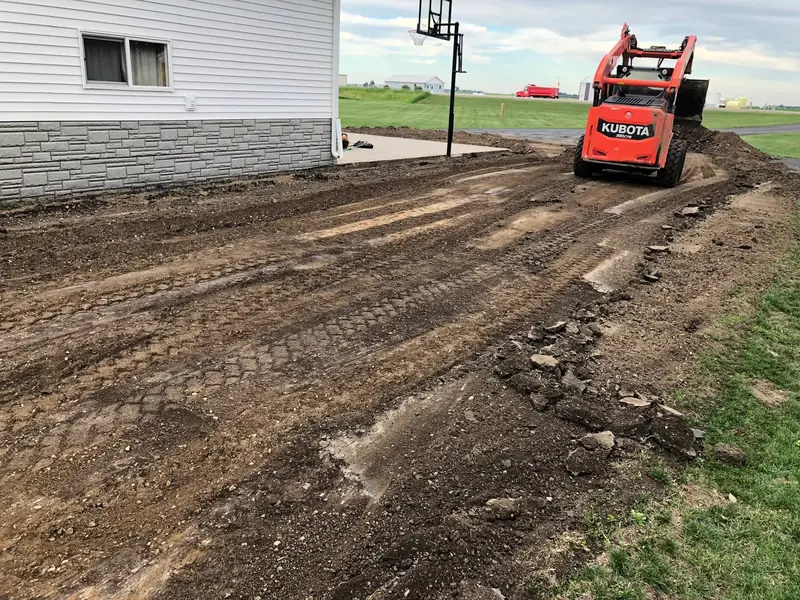 Skid steer excavating and grading a driveway route beside a house before paving