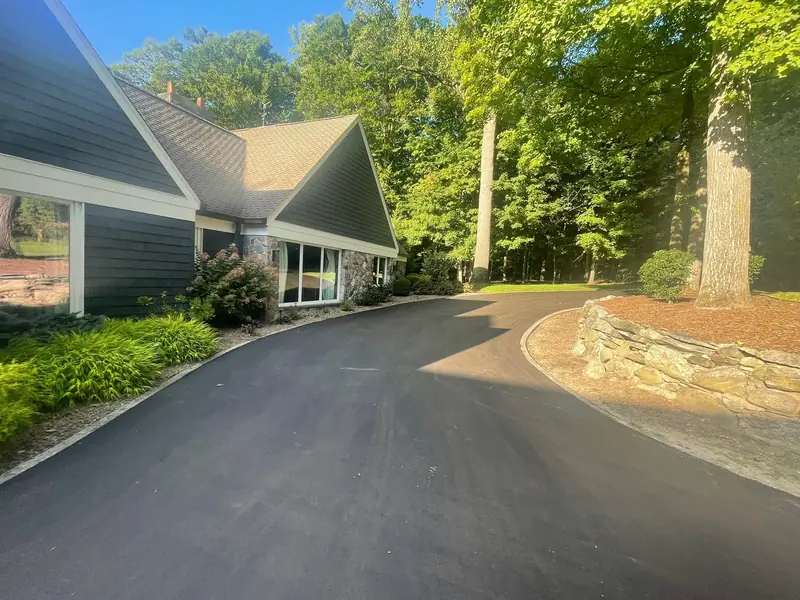 Freshly paved driveway curving past a stacked-stone wall and landscaped beds to a wooded home